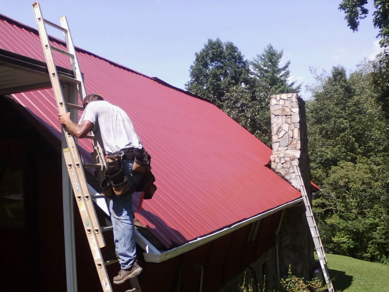 Roofer on a ladder beside a red metal roof and stone chimney — Soffit & Fascia Repair by Roofing Brothers in North Garden